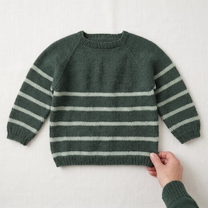 May include: A long-sleeve knit jumper featuring a dark forest green base with horizontal light sage green stripes. The garment has a round crew neckline, ribbed cuffs, and a ribbed hem, constructed with a classic stockinette stitch pattern.