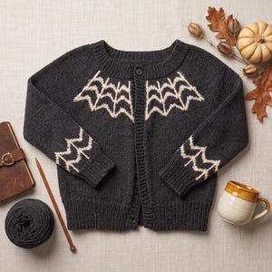 May include: A dark charcoal gray knit cardigan featuring a cream-colored spiderweb pattern across the yoke and cuffs. The garment has a round neckline, a button-front closure, and ribbed trim along the collar, cuffs, and hemline.