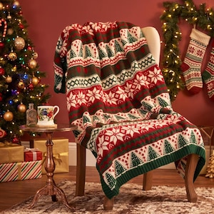 Pode incluir: Uma manta de malha com um padrão festivo de pinheiros verdes, flocos de neve brancos e formas geométricas em vermelho, branco e verde escuro. A manta está sobre uma cadeira, exibindo um design repetitivo com tema de inverno.