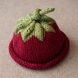 Può includere: Un berretto lavorato a maglia a forma di fragola, caratterizzato da un corpo rosso scuro con bordo arrotolato e una sezione superiore verde chiaro che imita le foglie, completo di un piccolo stelo annodato sulla sommità per una finitura.