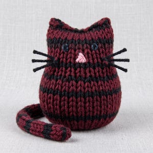 Puede incluir: Una pequeña figura de gato tejida a mano con rayas horizontales en lana color burdeos oscuro y negro. El juguete de peluche tiene dos ojos bordados en azul oscuro, una pequeña nariz cosida en rosa claro y bigotes de lana negra.