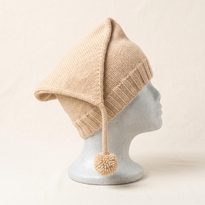 May include: A beige knit hat featuring a pointed crown and a ribbed brim. The hat has a long, triangular flap extending from the top, which hangs down the side and terminates in a small, round pom-pom made of matching beige yarn.