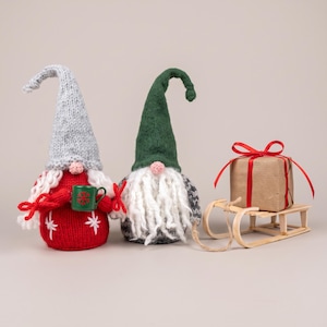 May include: Two plush gnomes with long white beards and tall pointed hats. One gnome wears a grey hat and red jumper with white snowflake details, holding a green mug. The other wears a dark green hat and grey patterned jumper. A wooden sledge carries a brown gift box with a red ribbon.