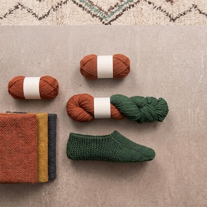 Könnte beinhalten: Zwei rostfarbene Wollknäuel, ein Wollstrang mit Farbverlauf von Rost zu Dunkelgrün, ein dunkelgrüner Strickhausschuh und ein Stapel aus drei gefalteten Strickmustern in den Farben Rost, Senfgelb und Dunkelgrau.