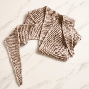 Puede incluir: Una bufanda de punto de color beige claro que presenta una textura acanalada distintiva en toda su longitud. La bufanda tiene una forma alargada y cónica con extremos puntiagudos y un patrón de puntadas uniforme que crea un diseño lineal sutil.