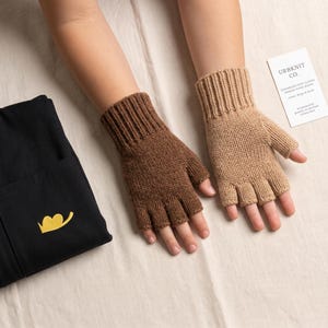 May include: A pair of fingerless knit gloves in brown and beige, featuring a ribbed cuff and open fingers. Beside them lies a black fabric item with a yellow embroidered heart-shaped logo and a white product tag labeled URBKNIT CO.