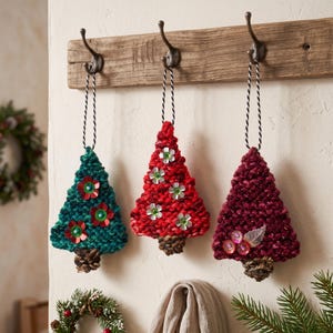May include: Three triangular Christmas tree ornaments made of chunky knit yarn in teal, bright red, and deep burgundy. Each ornament features a small pinecone base, decorative sequins or metallic leaf accents, and a black and white twisted cord loop.