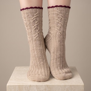 Könnte beinhalten: Ein Paar beigefarbene Strickstrümpfe mit einem vertikalen Zopfmuster an der Vorderseite und den Seiten. Die Socken haben eine gerippte Textur am gesamten Bein und Fuß und sind mit einem dunkelroten, gewellten Rand am oberen Bündchen versehen.