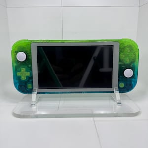 May include: A handheld gaming console featuring a translucent shell that transitions from a vibrant lime green at the top to a deep teal blue at the bottom. The device includes a central black screen, white analog sticks, and green directional buttons.