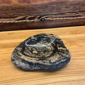 May include: A natural stone bowl with a rough, textured exterior and a smooth, polished interior basin. The stone features a mottled pattern of black, gray, and white mineral deposits, creating a rugged appearance with a glossy, reflective center.