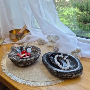 Op de afbeelding: Twee natuurstenen schalen met ruwe, getextureerde buitenkanten en gepolijste, gladde binnenkanten. De ene schaal is gespikkeld grijs en bruin en bevat rode en witte sieraden. De andere is donkergrijs met witte aders en bevat twee kralenarmbanden.