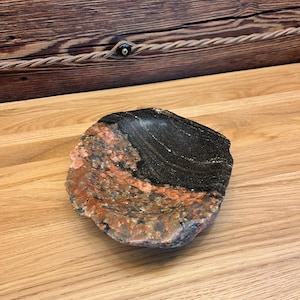 Op de afbeelding: Een ondiepe, ronde stenen kom met een natuurlijke, ruw afgewerkte buitenrand. Het binnenoppervlak vertoont een opvallend contrast tussen een gespikkeld gedeelte met oranje, grijze en zwarte mineralen en een glad, donkerzwart gedeelte.