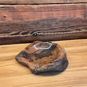 May include: A polished stone bowl featuring a natural, irregular shape with a mottled pattern of orange, brown, and black granite. The interior is smooth and concave, while the exterior retains a rugged, raw texture with visible mineral grains.