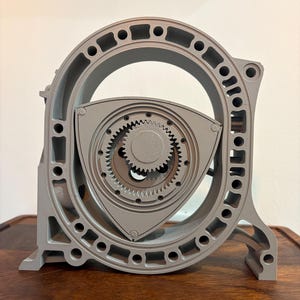 May include: A grey 3D printed model of a Wankel rotary engine housing and rotor. The assembly features a triangular rotor with a central gear positioned inside a stationary oval-shaped housing with multiple circular bolt holes and structural cutouts.