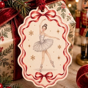 May include: A decorative hanging ornament featuring a scalloped edge with a red ribbon border. The centre illustration depicts a ballerina in a white tutu and pointe shoes, surrounded by gold-coloured snowflakes and stars on a cream background.