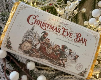 Könnte beinhalten: Eine rechteckige Milchschokoladentafel mit zartem Karamell in einer weißen Verpackung mit dem Text Mrs. Lamb's Sweet Emporium Christmas Eve Bar. Das Etikett zeigt Elfen, die einen roten Schlitten in einer verschneiten Waldlandschaft beladen.