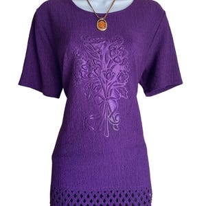 May include: A short-sleeved purple top with a textured fabric surface, featuring a central embossed floral bouquet design and a decorative lattice-style cutout hem. A brown cord necklace with a round amber-colored pendant rests at the neckline.