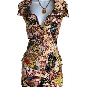 May include: A sleeveless button-down top featuring a vibrant butterfly print in shades of yellow, red, black, and white. The garment has a wide collar, a front button closure, and a fitted silhouette. An amber-colored pendant hangs on a brown cord.
