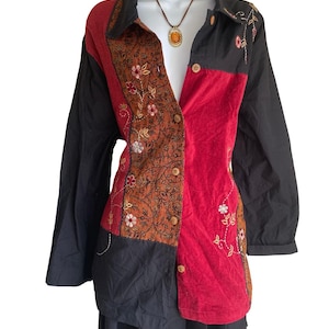 May include: A long-sleeved button-down shirt featuring a color-blocked design in black and red, with a central panel of rust-colored fabric adorned with intricate floral embroidery. The shirt includes a collar and light-colored wooden buttons.