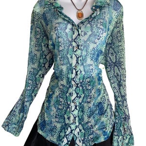 May include: A long-sleeved button-down shirt featuring a blue and green snakeskin pattern, paired with a black skirt. An oval amber-coloured pendant on a brown cord hangs around the neck area of the shirt, which is displayed on a white mannequin.