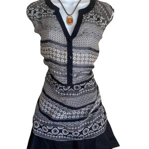 May include: A sleeveless tunic top featuring a black and white patterned print with horizontal bands of geometric and floral motifs. The top has a V-neckline with a black placket, three black buttons, and a solid black pleated hemline.