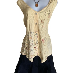 May include: A sleeveless cream-coloured blouse featuring a mix of polka dot and floral patterns with a front button closure, paired with a dark navy blue pleated skirt. A brown cord necklace with an oval amber-coloured pendant rests at the neckline.