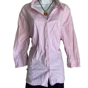 May include: A light pink and white gingham button-down shirt with long sleeves and a collar, displayed on a mannequin. A brown cord necklace with an oval amber-colored pendant rests around the collar area of the shirt over a black garment.