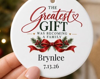 May include: A round white ceramic Christmas ornament featuring the red and green text THE GREATEST GIFT WAS BECOMING A FAMILY. Below the text is a red ribbon with green pine branches and red berries. The name Brynlee and date 7.13.26 are printed.