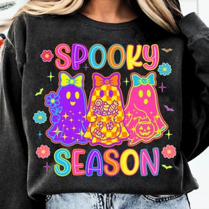 May include: A black crewneck sweatshirt featuring a colorful graphic with the text SPOOKY SEASON. The design includes three ghosts with bows, one holding a flower, one with a checkered pattern holding candy, and one holding a pumpkin bucket.