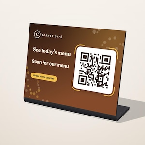 Puede incluir: Un letrero de acrílico marrón en forma de L con el logotipo de Corner Café, un código QR y el texto: See today's menu, Scan for our menu y Order at the counter. El letrero tiene un fondo degradado marrón oscuro con acentos circulares.