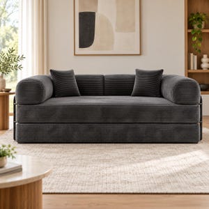 May include: A charcoal gray corduroy sofa featuring a low-profile design with rounded armrests and two matching square throw pillows. The upholstery has a distinct horizontal ribbed texture, and the frame is supported by a dark metal structure.