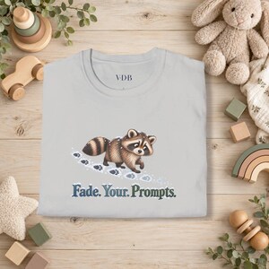 Peut inclure: Un t-shirt gris clair orné d'un motif représentant un raton laveur brun et noir marchant dans la neige, laissant des empreintes de pattes. Sous le raton laveur, le texte "Fade. Your. Prompts." est écrit en bleu foncé. Logo VDB au col.