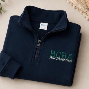 Op de afbeelding: Een marineblauw sweatshirt met kwartritssluiting, opstaande kraag, lange mouwen met geribbelde boorden en een ritssluiting aan de voorzijde. Op de borst staat de groene geborduurde tekst BCBA boven de witte tekst Your Name Here.