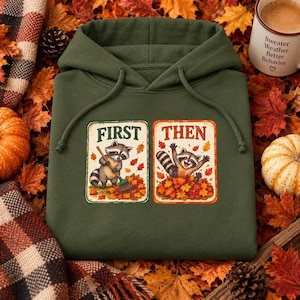 Op de afbeelding: Een olijfgroene hoodie met twee rechthoekige grafische prints. De linkerprint toont een wasbeer die herfstbladeren harkt met de tekst FIRST. De rechterprint toont een wasbeer die in een hoop bladeren speelt met de tekst THEN.