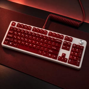 Könnte beinhalten: Eine mechanische Tastatur mit weißem Rahmen und durchscheinenden dunkelroten Tastenkappen mit weißer Beschriftung. Die Tastatur verfügt über einen vollständigen Ziffernblock, einen silbernen Drehknopf oben rechts und ein gewickeltes rotes Kabel.