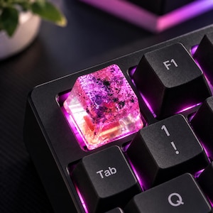 May include: A translucent, square mechanical keyboard keycap featuring a pink and purple glitter-infused resin design. The keycap is positioned on a black keyboard base, illuminated from beneath by a vibrant purple LED light, situated near the Tab key.