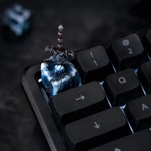 Könnte beinhalten: Eine individuelle mechanische Tastenkappe mit einem silbernen Miniaturschwert mit braunem Griff, das in einem strukturierten, eisblauen und weißen Felsensockel steckt und mit einer kleinen schwarzen Metallkette auf einer schwarzen Tastatur verziert ist.