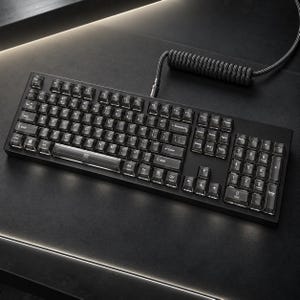 May include: A black mechanical keyboard featuring transparent keycaps with white lettering and a full numeric keypad. The keyboard is connected by a black coiled cable that extends from the top edge, showcasing a standard QWERTY layout.