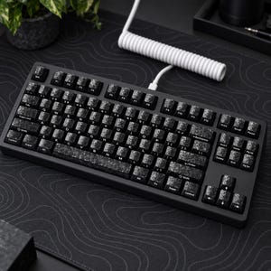 Könnte beinhalten: Eine mechanische Tastatur mit einem mattschwarzen Rahmen und Tastenkappen in einem dunklen Marmorsteinmuster. Die Tastatur verfügt über ein Standard-QWERTY-Layout mit weißer Beschriftung auf jeder Taste und ist über ein weißes Spiralkabel verbunden.
