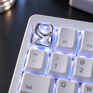 Puede incluir: Una tecla de teclado mecánico personalizada con un marco de metal de color plateado pulido con un diseño de corte orgánico e irregular. La tecla está instalada en un teclado blanco con retroiluminación LED azul brillante visible a través de la estructura.