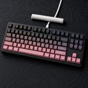 Puede incluir: Un teclado mecánico con marco negro y un conjunto de teclas con degradado de gris oscuro a rosa claro. Cada tecla presenta un patrón de líneas topográficas blancas. Un cable USB blanco en espiral está conectado a la parte superior del teclado.