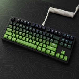 Könnte beinhalten: Eine mechanische Tastatur mit schwarzem Rahmen und einem Tastenkappenset mit Farbverlauf von Dunkelgrün zu Hellgrün. Die Tastatur verfügt über ein Standard-QWERTY-Layout mit schwarzen Funktionstasten und einem weißen Spiralkabel oben.