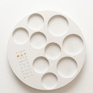 May include: A circular white concrete tray featuring eight recessed circular indentations of varying sizes. The surface includes a vertical column of Hebrew text engraved into the material, with the second line highlighted in gold paint.