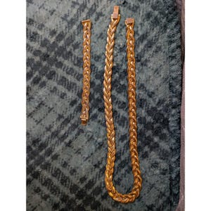 May include: A gold-toned metal jewellery set consisting of a shorter braided chain bracelet and a longer braided chain necklace. Both pieces feature a consistent woven texture and a rectangular clasp mechanism at the ends of each chain segment.