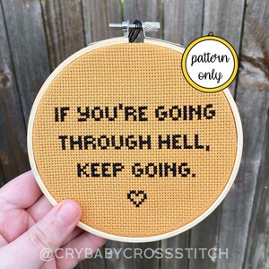 May include: A yellow cross-stitch hoop with the text "IF YOU'RE GOING THROUGH HELL, KEEP GOING." in black thread. A small heart is stitched below the text. The hoop is held in a hand. The text "pattern only" is written on a yellow circle in the upper right corner of the image.