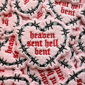 May include: Pink fabric patch with a black barbed wire border and red embroidered text that reads "heaven sent hell bent".