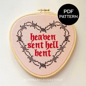 May include: A pink and white cross-stitch embroidery in a wooden hoop, featuring the text "heaven sent hell bent" in red and black gothic lettering, surrounded by a black barbed wire design.
