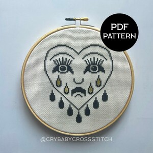 May include: A black and white cross stitch pattern of a crying heart with eyelashes, a sad mouth, and teardrop-shaped beads. The pattern is framed in a wooden hoop and the text "@CRYBABYCROSSTITCH" is at the bottom. The text "PDF PATTERN" is in a black circle at the top right of the image.