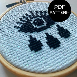 May include: A black and white cross stitch pattern of a pixelated eye with the text "PDF PATTERN" in a black circle above it. The cross stitch is framed in a wooden hoop.