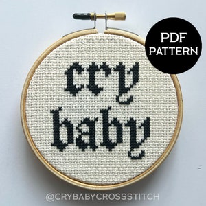 May include: A black and white cross stitch pattern in a wooden embroidery hoop, featuring the text "cry baby" in a gothic font. The pattern is available as a PDF download.